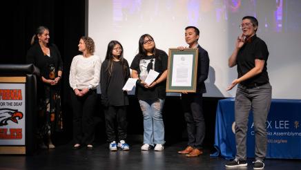 Asm. Lee holding a plaque with students and interpreters on stage