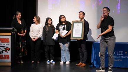 Asm. Lee holding a plaque with students and interpreters on stage