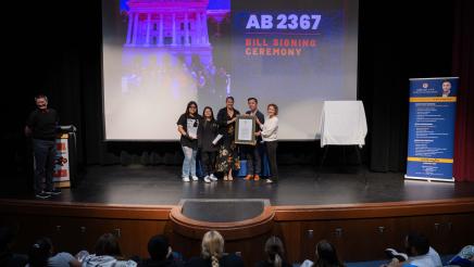 Wide photo of Asm. Lee holding a plaque with students and interpreters on stage